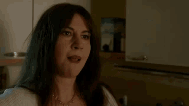 a woman with long dark hair is sitting in a kitchen with a necklace on her neck .