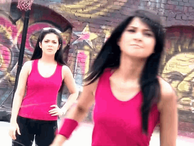 two women in pink tank tops stand in front of a brick wall with graffiti on it