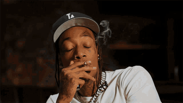 a man wearing a hat with the letter t on it smoking a cigarette