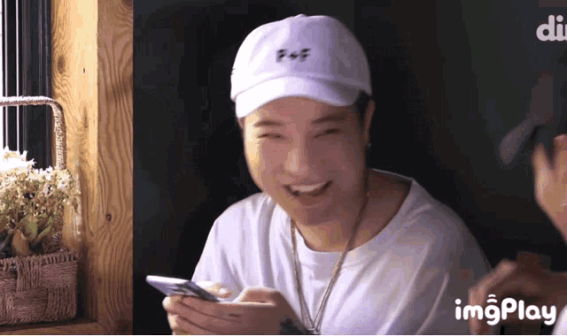 a man wearing a white hat and a white t-shirt is smiling and holding a cell phone