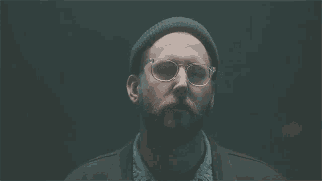 a man with a beard wearing glasses and a beanie looks at the camera