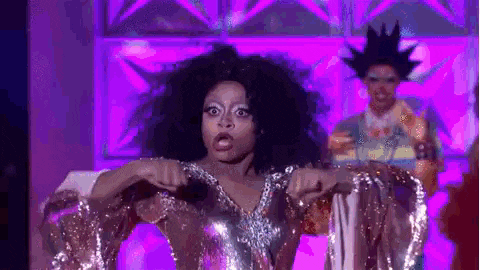 a woman in a silver dress is dancing on a stage with a man in a wig behind her .