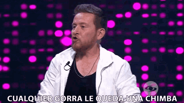 a man in a white jacket is talking into a microphone with the words " cualquier gorra le queda una chimba " above him