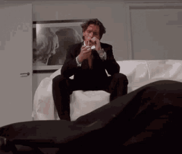 a man in a suit is sitting on a white couch smoking a cigarette .