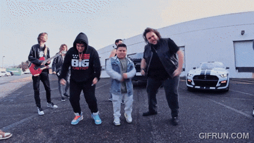 a group of people are dancing in a parking lot and one of them is wearing a shirt that says big