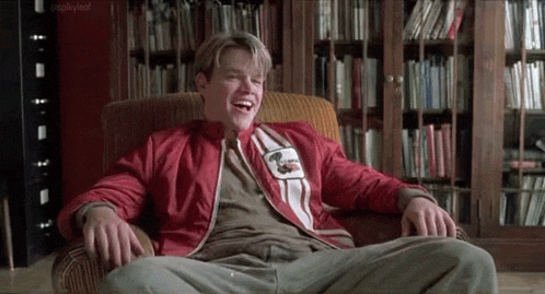 a man in a red jacket is sitting in a chair in front of a bookshelf and laughing .
