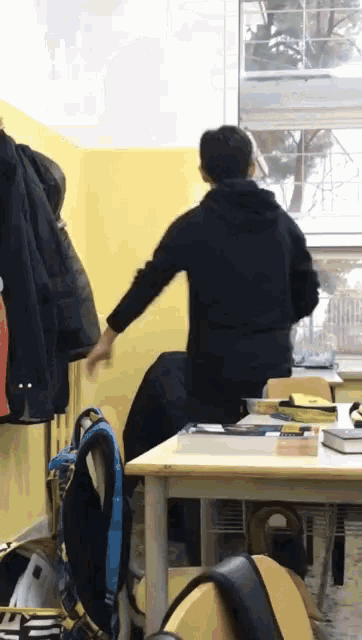 a man in a black hoodie stands in a classroom with his arms outstretched