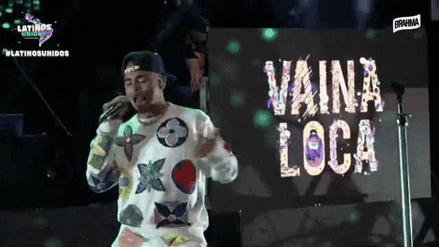 a man singing into a microphone with the words vaina loca on the screen behind him