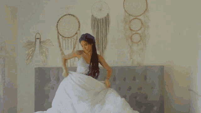 a woman is sitting on a bed with a white blanket and a dream catcher on the wall .