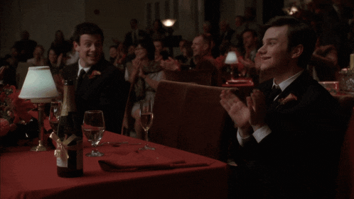 a man in a tuxedo sits at a table clapping his hands