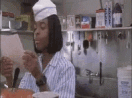 a woman in a chef 's hat is sitting in a kitchen eating a sandwich .