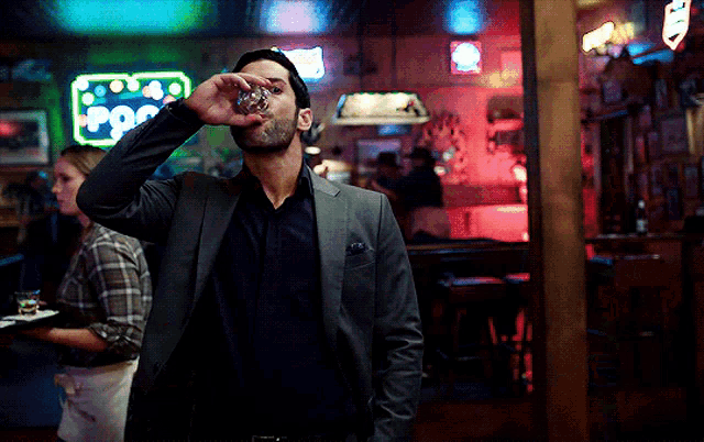 a man in a suit drinks from a shot glass in front of a neon sign that says ' pop '