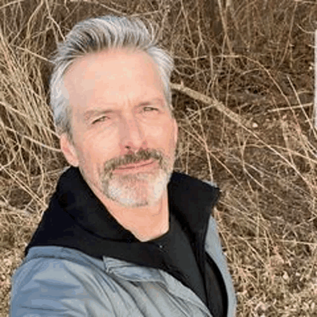 a man with gray hair and a beard is taking a selfie in a field .