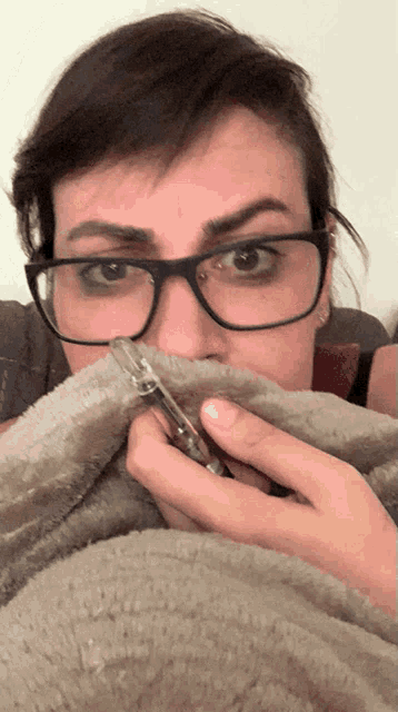 a woman with glasses covering her mouth with a blanket while holding a cell phone