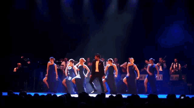 a man in a tuxedo stands on a stage with a group of dancers