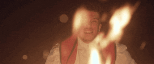 a man in a white shirt and red vest is standing in front of a fire in the dark .
