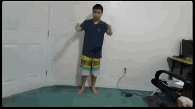 a young boy in a blue shirt and yellow shorts is standing on a green rug in a room .