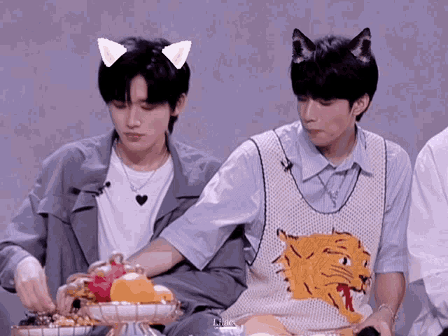 a man wearing a sweater vest with a tiger on it sits next to another man wearing cat ears