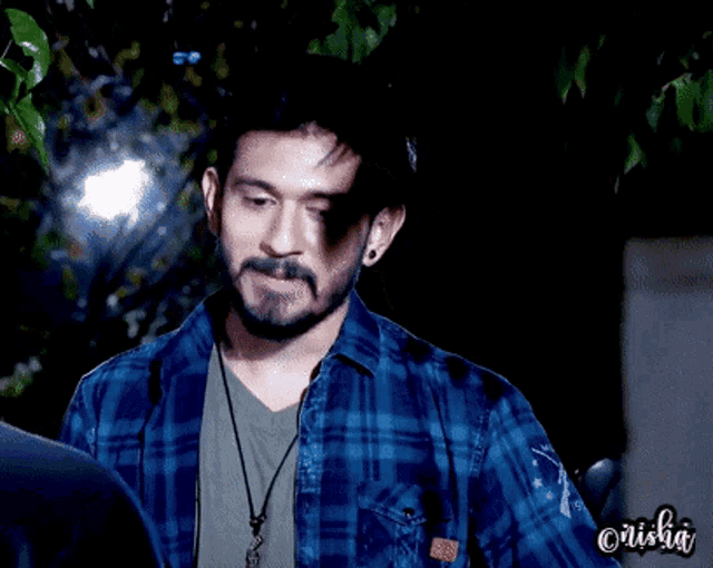 a man wearing a blue plaid shirt and a necklace with the name oiisha on the bottom right