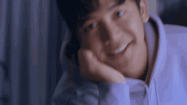 a young man wearing a purple hoodie is smiling with his hand on his face .