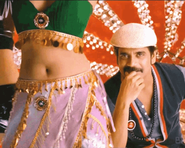 a man with a mustache is covering his mouth while a woman in a green top and purple skirt stands behind him