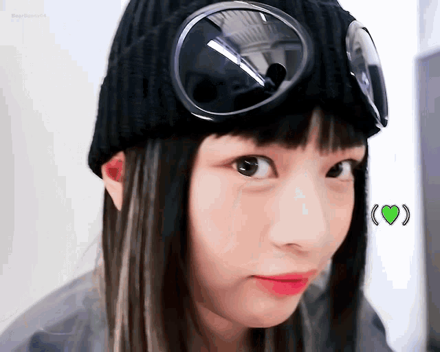 a girl wearing a black beanie and goggles with a green heart in the background .