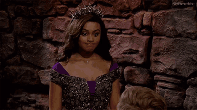 a woman in a purple dress with a tiara on her head stands in front of a stone wall