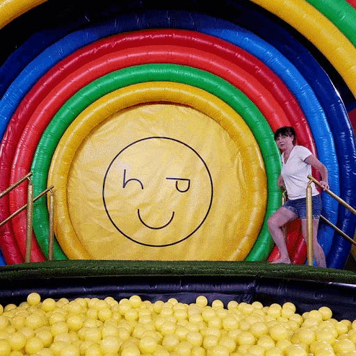 a woman stands in front of a large inflatable rainbow with a smiley face drawn on it
