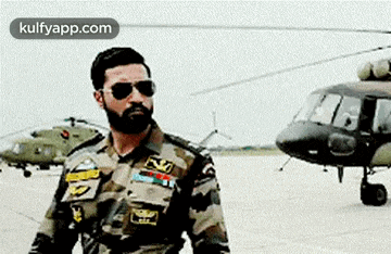 a man in a military uniform and sunglasses is standing in front of a helicopter on a runway .