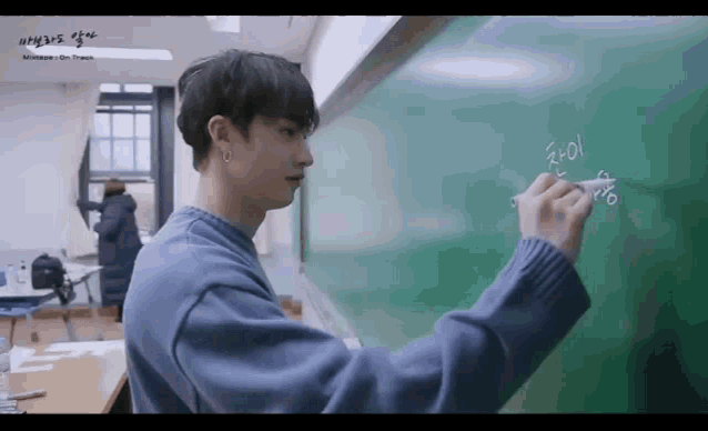 a man in a blue sweater is writing on a blackboard in a classroom .