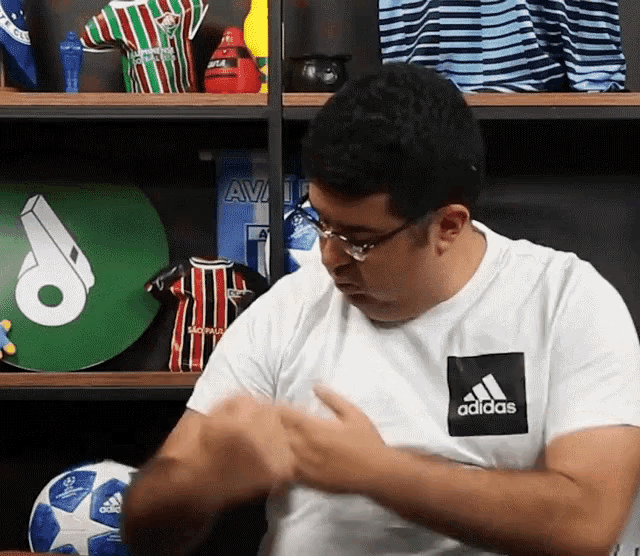 a man wearing a white adidas t-shirt is sitting in front of a shelf full of soccer jerseys