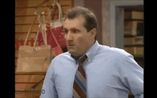 a man in a blue shirt and tie is standing in front of a wall of handbags .