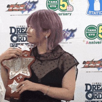 a woman with purple hair holds a wrestling belt in front of a wall that says tama kyu