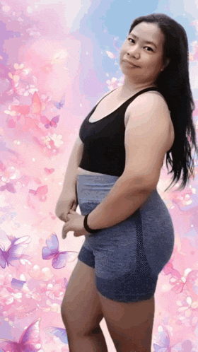 a woman in a black tank top and blue shorts stands in front of a pink background with butterflies .