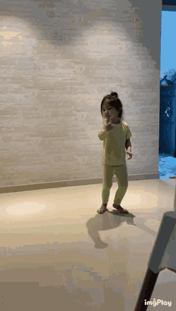 a little girl in a yellow shirt and green pants is standing on a tiled floor ..
