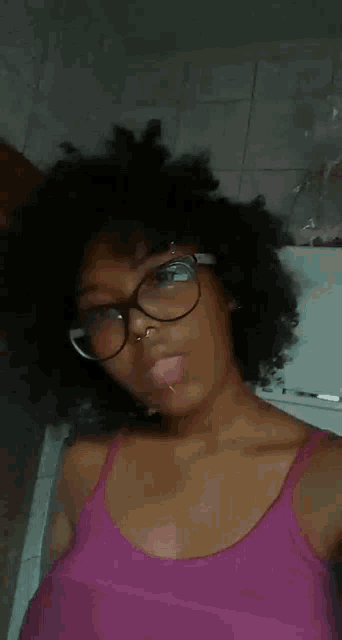 a woman wearing glasses and a pink tank top has her tongue sticking out