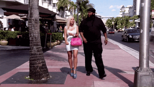 a woman in a white dress walks down a sidewalk next to a man in a black suit
