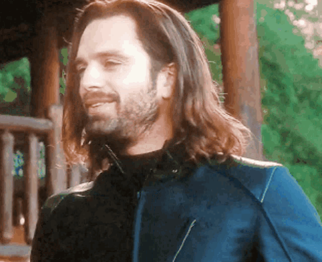 a man with long hair and a beard is standing in front of a wooden structure .