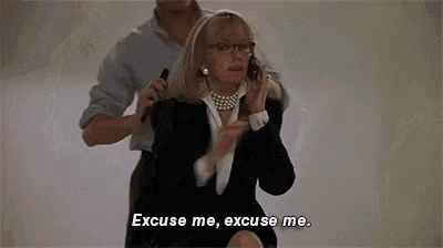 a woman in a suit is talking on a cell phone and a man is standing behind her .