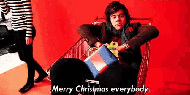harry styles is sitting in a shopping cart with a gift box on his lap and says merry christmas everybody .