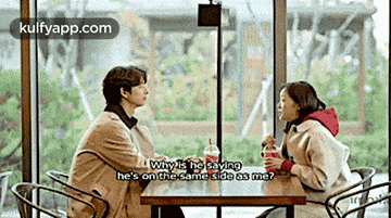 a man and a woman are sitting at a table in front of a window talking to each other .