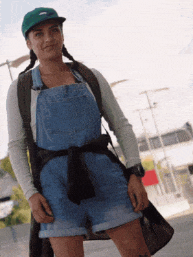 a woman wearing overalls and a green hat has a backpack