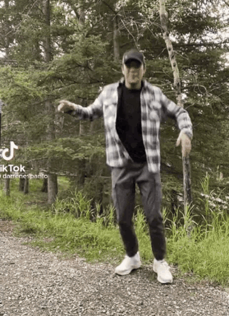 a man in a plaid shirt and hat is dancing on a gravel road in the woods .