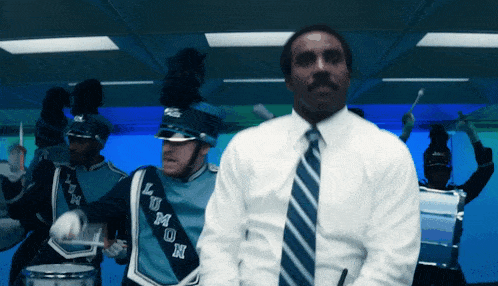 a man in a white shirt and tie stands in front of a marching band wearing blue uniforms that say lum on them