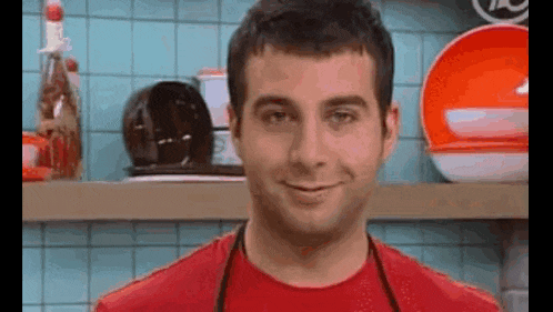 a man in a red shirt and apron is standing in a kitchen .