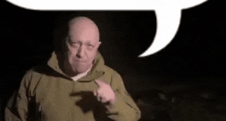 a bald man in a green jacket is pointing at the camera .