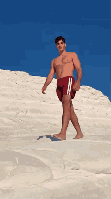 a shirtless man in red swim trunks stands on a white sandy hill