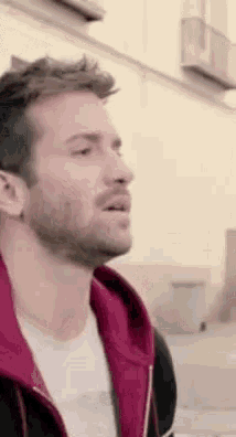 a man with a beard wearing a red hoodie is standing in front of a building .