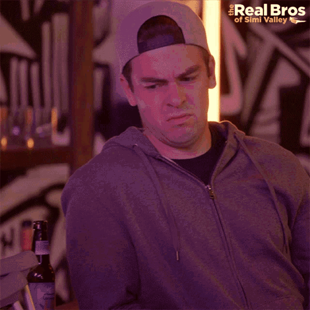 a man wearing a baseball cap and a grey hoodie with the real bros of simli valley written on the bottom