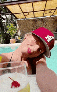 a woman is taking a selfie in a swimming pool while wearing a hat and holding a drink .